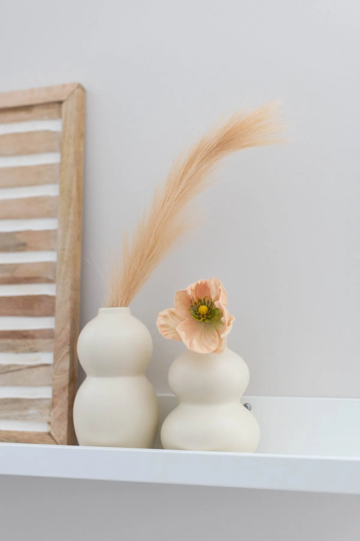 Two pale ceramic vases holding a dried grass stem and a peach bloom on a white picture ledge.